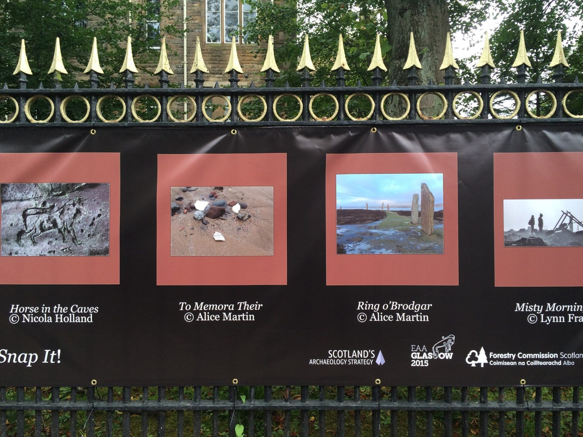Large black banner on a black and gold iron fence with two archaeological photographs, one with fragments on a beach titled 'To Memora Their' and another of standing stones in Orkney named 'Ring o'Brodgar', both with a red border.