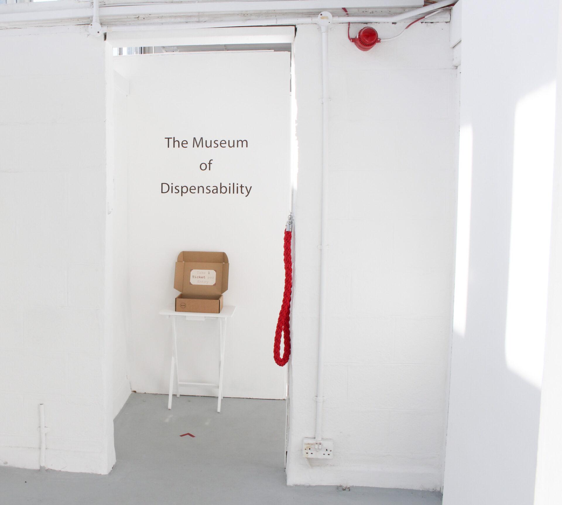 A white door frame with a red rope on the right, featuring vinyl text with the words 'The Museum of Dispensability' on the wall above a brown box on a white table.