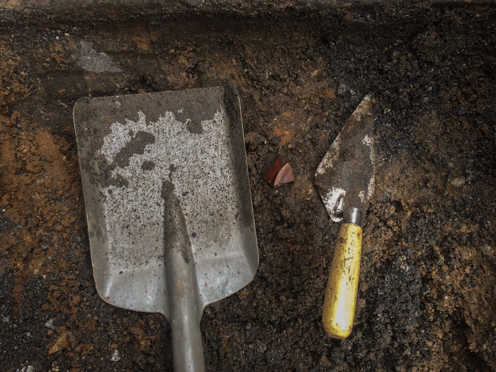A piece of red ceramics in between a small shovel on the left and a trowel on the right.