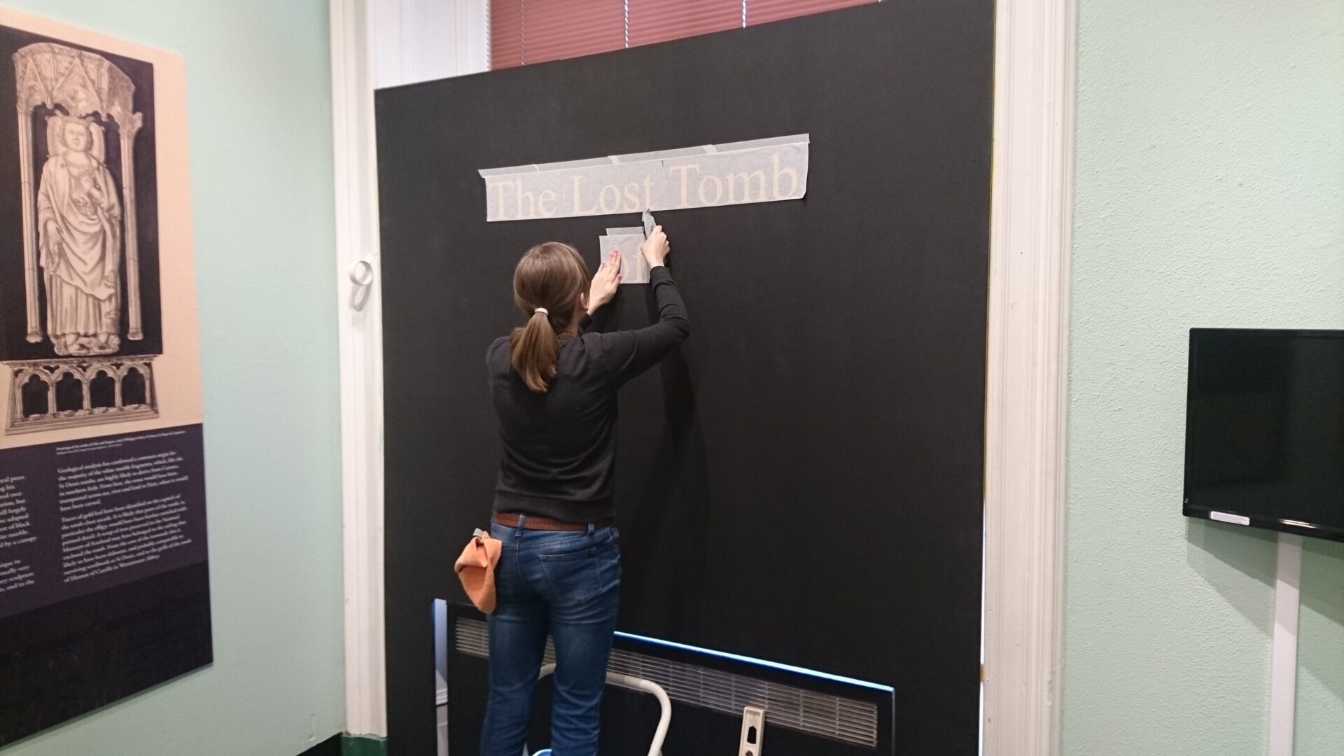 Artist Alice Martin sticking vinyl text reading 'The Lost Tomb' to a black wall.