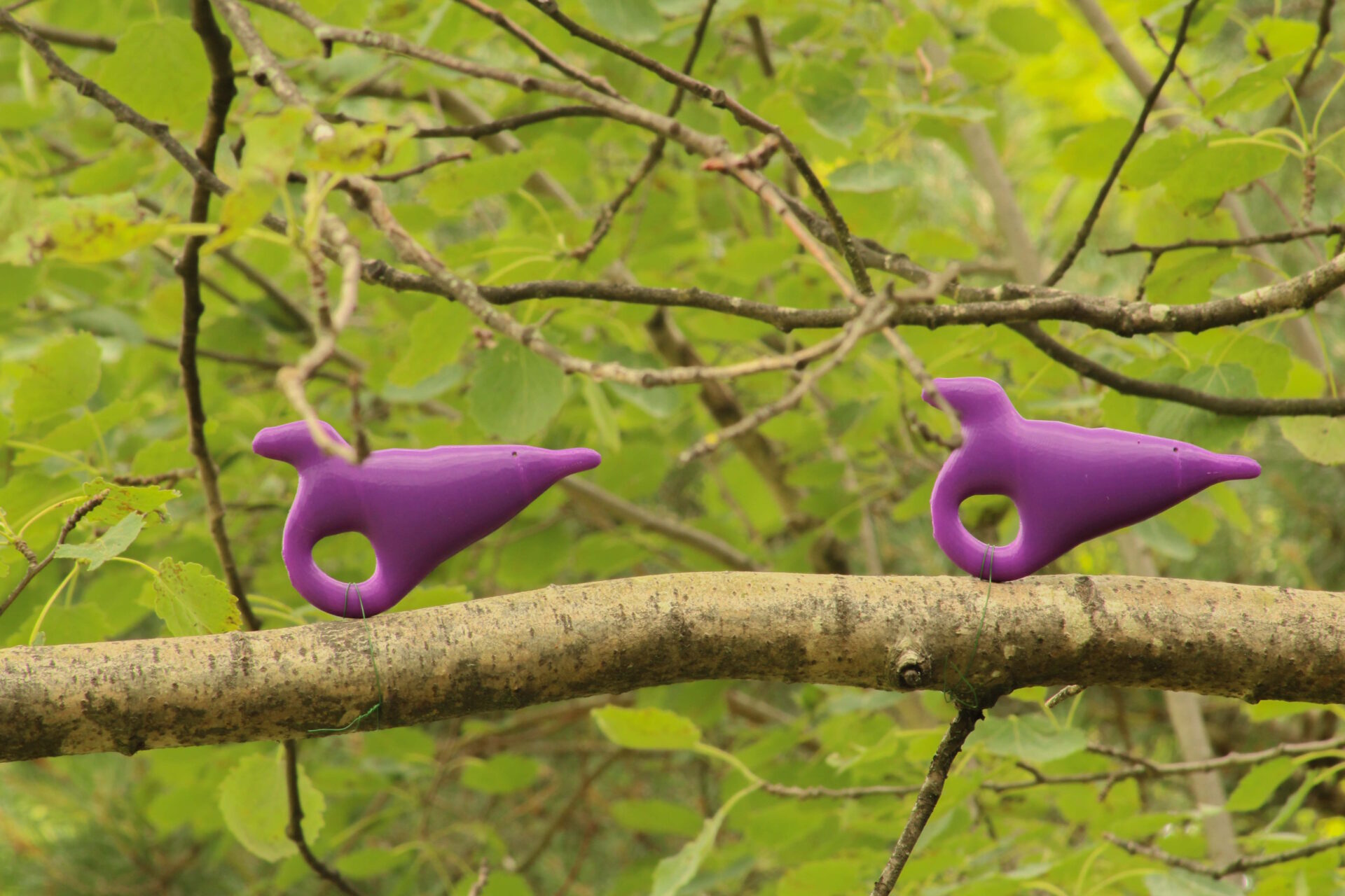 Two purple 3D prints resembling abstract birds perched on a branch with green leaves in the background.