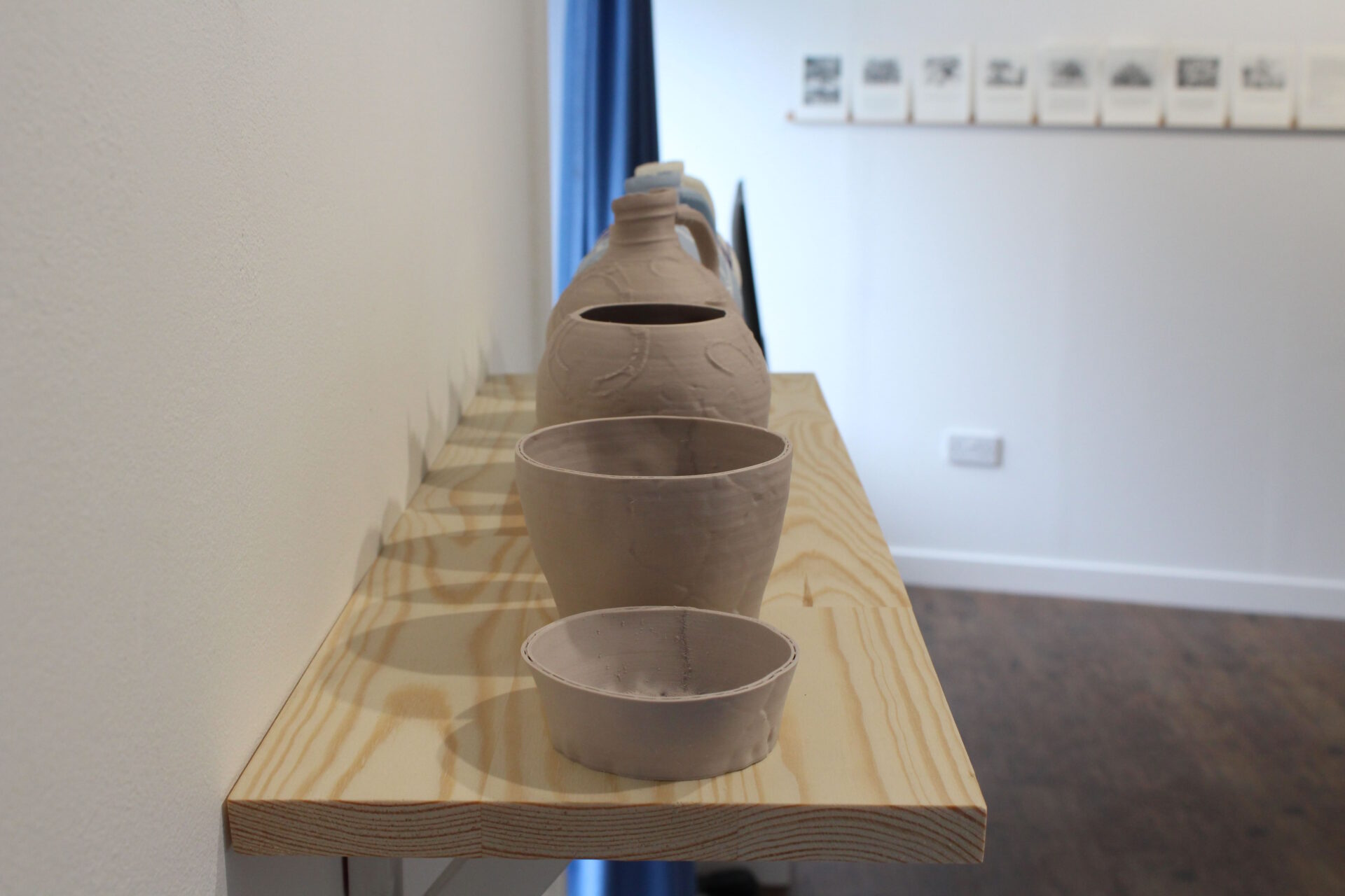 A series of grey clay 3D printed jugs in varying heights on a wooden shelf in a white gallery space.