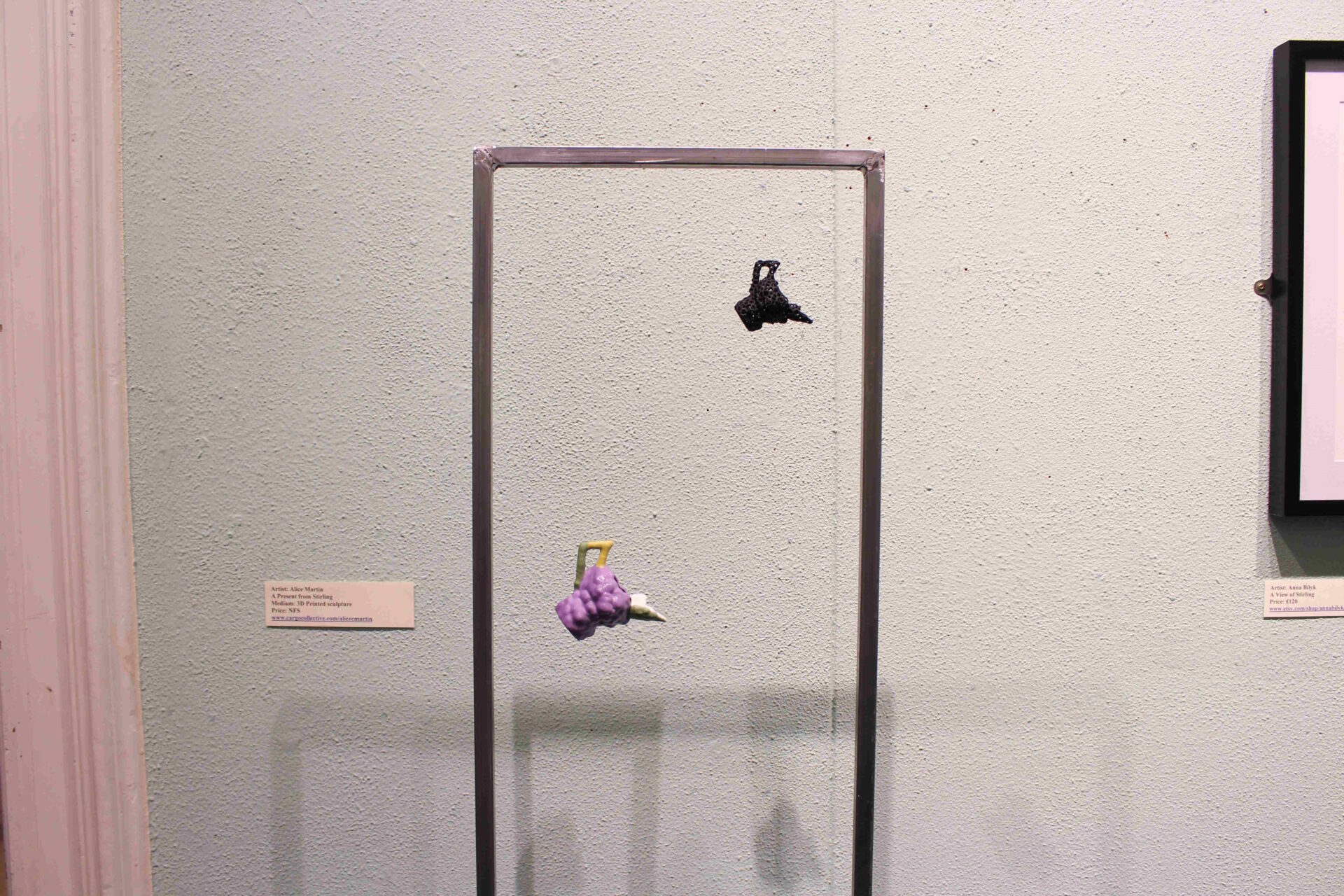 A purple, green and gold 3D printed jug and a black rubber one suspended from a metal frame using transparent fishing wire.