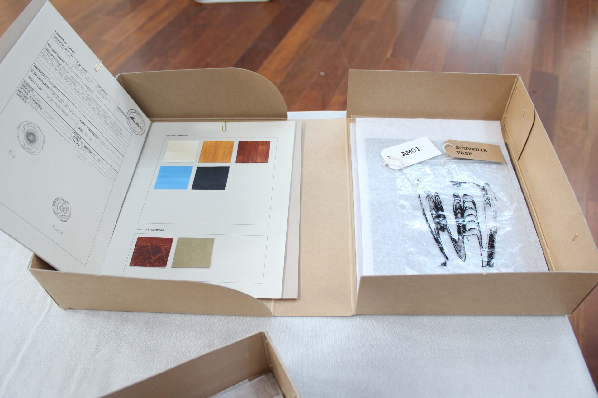 Archive box open with a file describing a vase with text, pencil drawings, colour, texture samples in brown, blue and black paint, white and brown labels and a black print inside a finds bag.