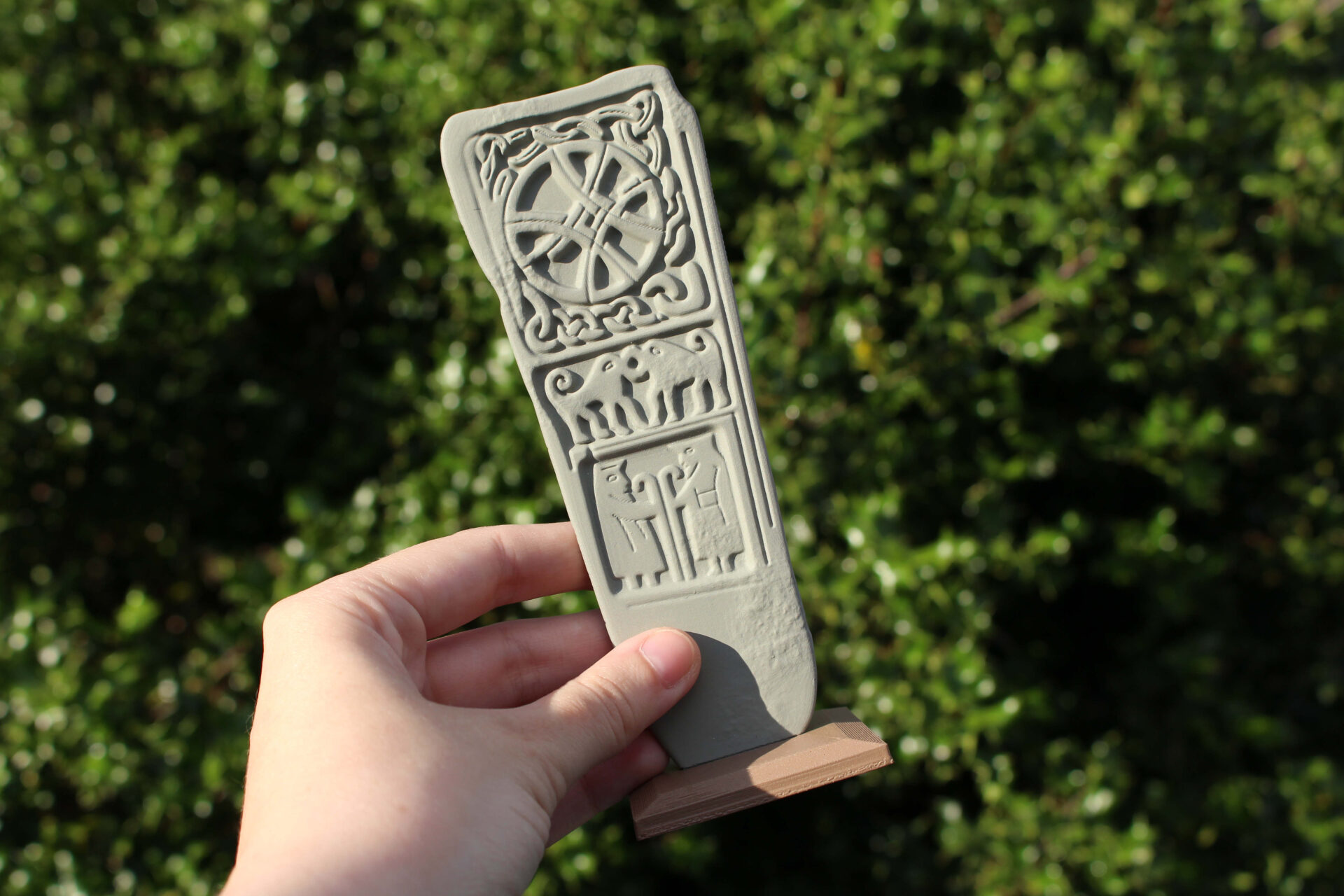 A hand holding a grey Pictish stone with symbols including a circle, interlace, two animals and bishops on a brown wooden base with greenery in the background.